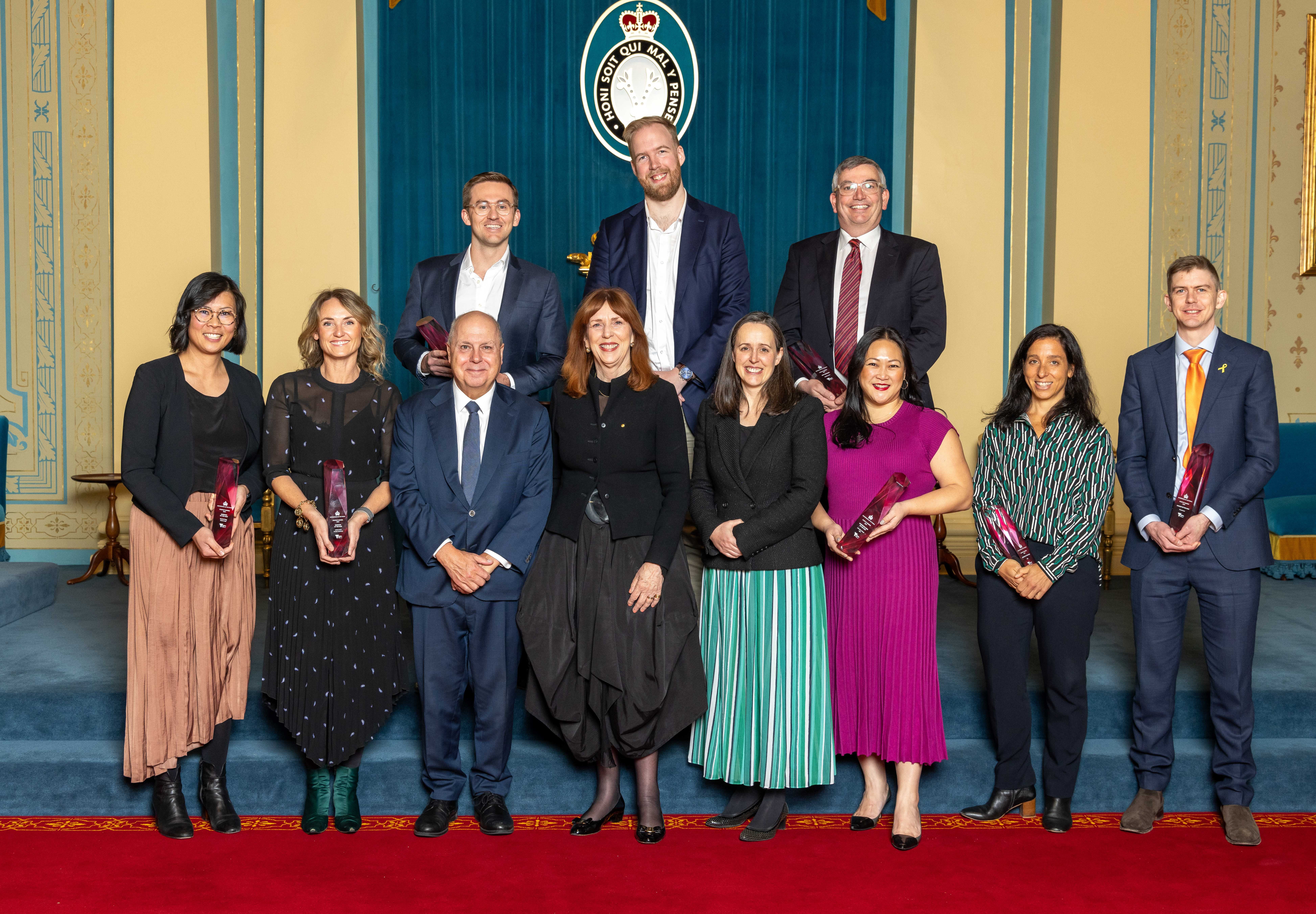 Winners of the awards stand with their awards in hand for a formal photo. There are two rows of smiling faces upon the small set of stairs. 